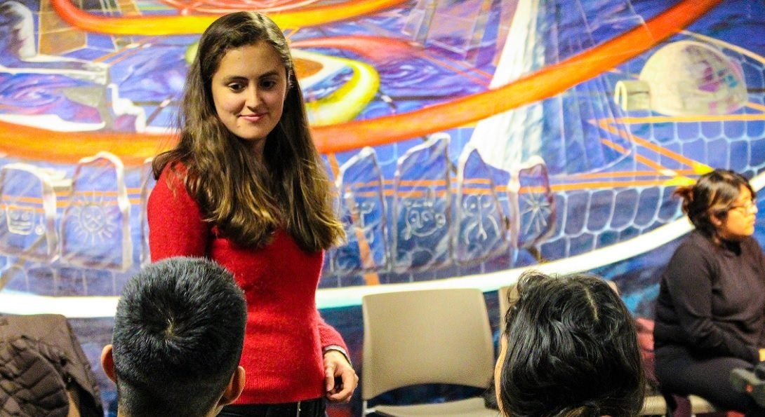 Latina listening to students, has faint smile. Colorful mural in the background.