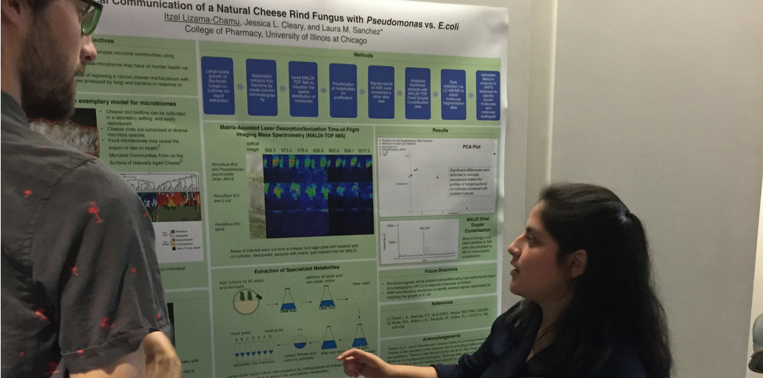 Latina scientist in dark shirt pointing to her scientific poster as man with a beard looks on