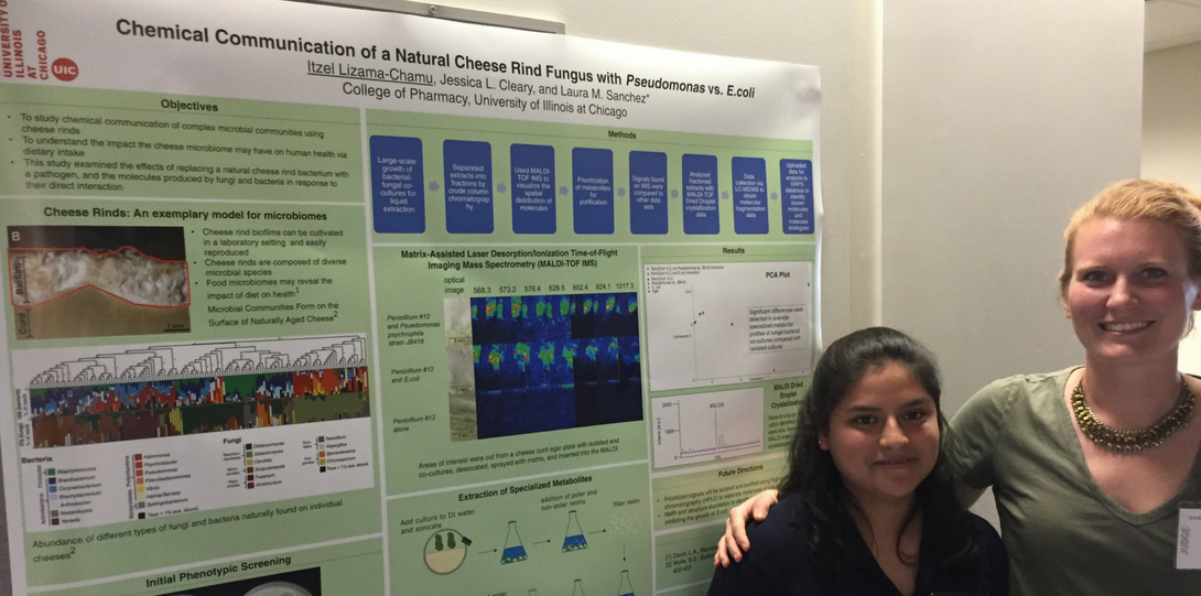Latina scientist in dark shirt stands in front of her scientific poster as a fair-skinned woman in olive shirt embraces her.