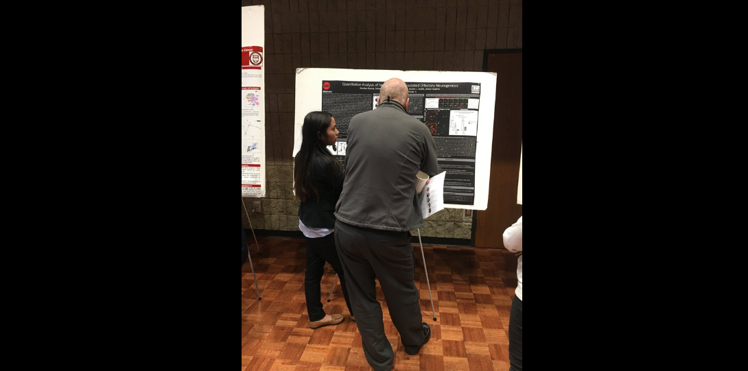 Hispanic female, Jocelyn, opposing the frame and facing a black poster that contains a synopsis of her research. Jocelyn is showcasing her research to an older white male, that's also opposing the frame and facing the poster, standing with a lean on his left hip.