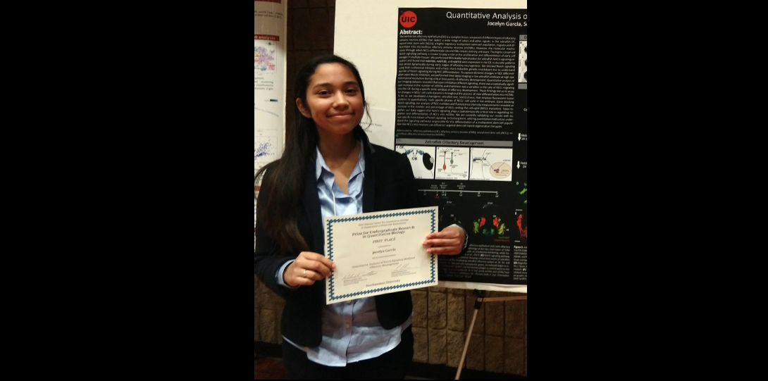 Hispanic female, Jocelyn, maintaining a cheerful grin whilst holding her first place award near her mid section with both hands. Student standing adjacent to her black poster that contains a synopsis of her research.