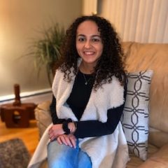 woman with curly hair sitting in a cream couch wearing jeans, a black long sleeve with her arms crossed and a tan cardigan over her long sleeve.